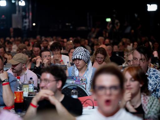 Ein Delegierter auf dem Parteitag der Linken sitzt unter Parteigenossen und hat ein Palästinensertuch als Symbol der Solidarität mit Palästina um seinen Kopf gebunden. Ein Delegierter auf dem Parteitag der Linken sitzt unter Parteigenossen und hat ein Palästinensertuch als Symbol der Solidarität mit Palästina um seinen Kopf gebunden.