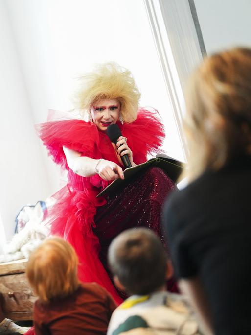 Die Drag Queen Freya Van Kant liest in einem roten Rüschenkleid und mit voluminöser blonder Frisur vor einer Kindergruppe aus einem Buch vor.