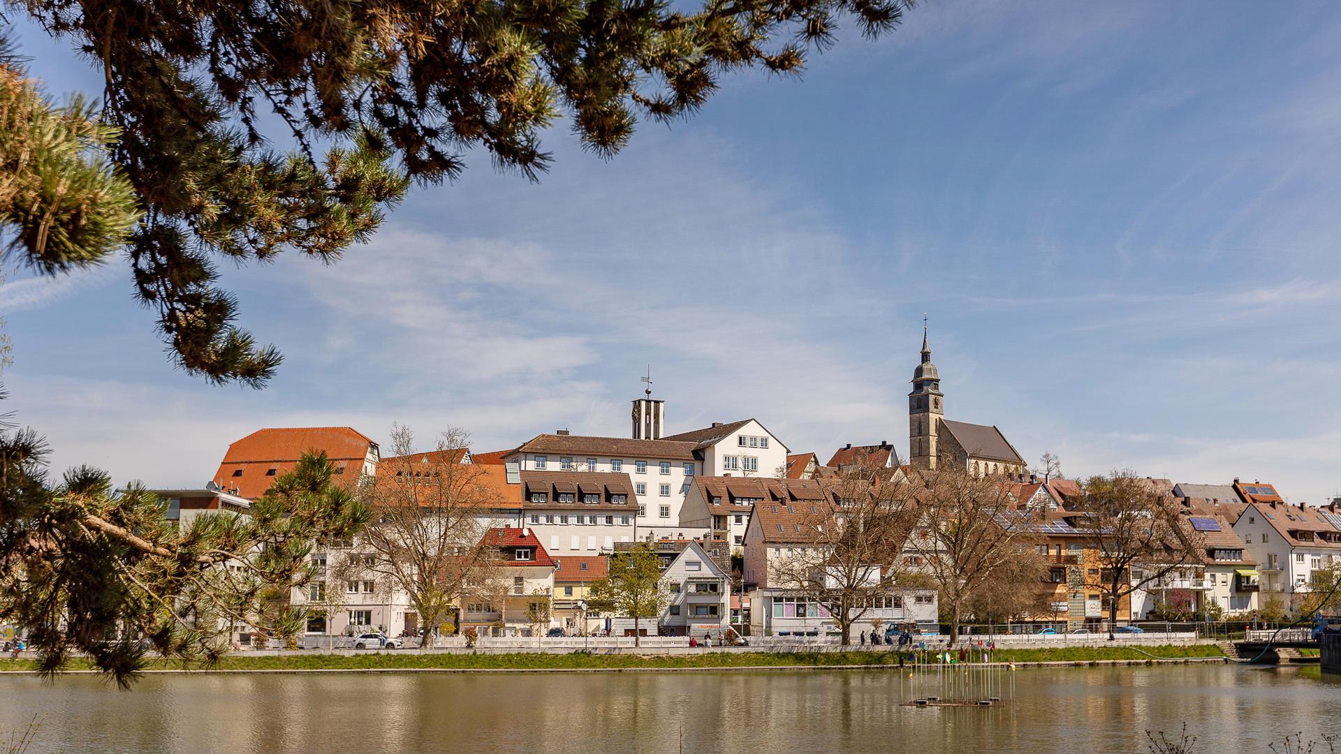 Stadtansicht von Böblingen mit blauem Himmel. Im Bildvordergrund ist der Untere See zu sehen, vom linken Bildrand ragt ein großer Ast eines Nadelbaums ins Bild.