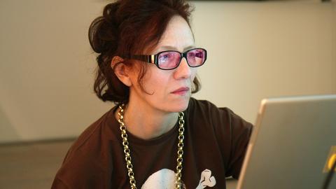 Cornelia Sollfrank sitzt an einem Schreibtisch und schaut konzentriert auf ihren Laptop. Sie trägt eine Brille mit rosafarbenen Gläsern und eine Goldkette um den Hals.