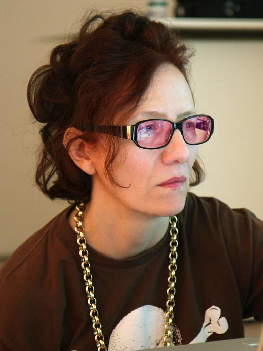 Cornelia Sollfrank sitzt an einem Schreibtisch und schaut konzentriert auf ihren Laptop. Sie trägt eine Brille mit rosafarbenen Gläsern und eine Goldkette um den Hals.