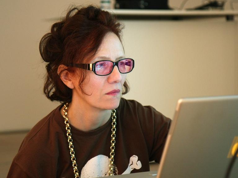 Cornelia Sollfrank sitzt an einem Schreibtisch und schaut konzentriert auf ihren Laptop. Sie trägt eine Brille mit rosafarbenen Gläsern und eine Goldkette um den Hals.