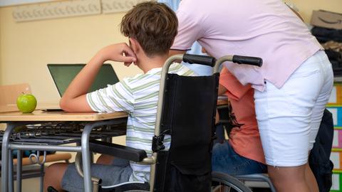 Ein Jugendlicher im Rollstuhl sitzt in einem Klassenzimmer am Tisch an einem Laptop. Neben steht eine erwachsene Person, die ihm assistiert. Die Personen sind von hinten zu sehen und somit nicht zu erkennen.