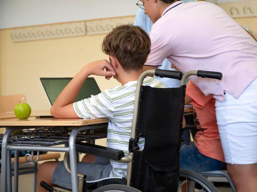 Ein Jugendlicher im Rollstuhl sitzt in einem Klassenzimmer am Tisch an einem Laptop. Neben steht eine erwachsene Person, die ihm assistiert. Die Personen sind von hinten zu sehen und somit nicht zu erkennen.