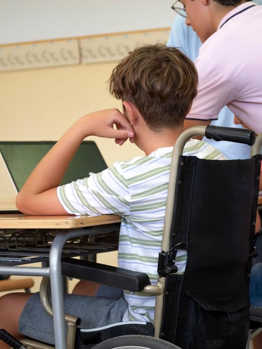 Ein Jugendlicher im Rollstuhl sitzt in einem Klassenzimmer am Tisch an einem Laptop. Neben steht eine erwachsene Person, die ihm assistiert. Die Personen sind von hinten zu sehen und somit nicht zu erkennen.