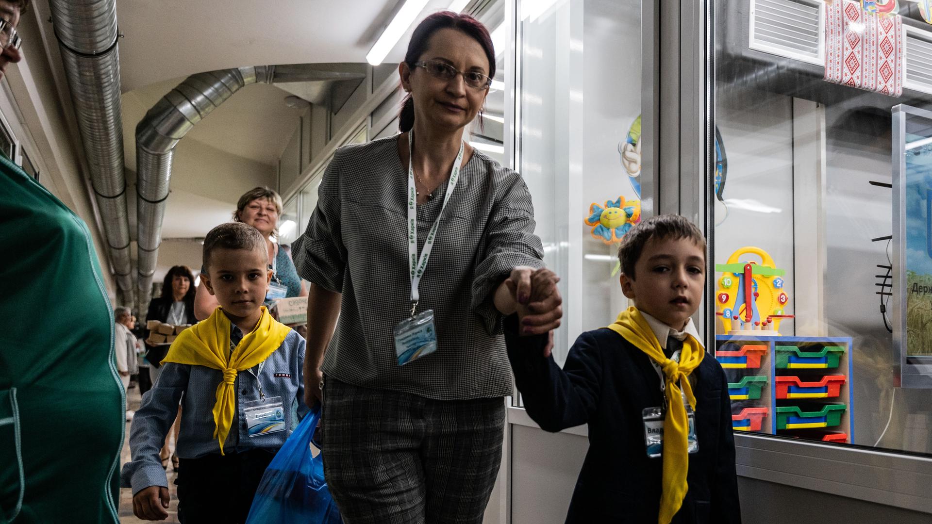 Schulbeginn in einer Metro-Station in Charkiw, im Osten der Ukraine. Eine Frau führt zwei Kinder an der Hand an Räumen vorbei, in die man durch große Glasfenster sehen kann. An der Decke sind dicke Lüftungsrohre zu sehen. Schulbeginn in einer Metro-Station in Charkiw, im Osten der Ukraine. Eine Frau führt zwei Kinder an der Hand an Räumen vorbei, in die man durch große Glasfenster sehen kann. An der Decke sind dicke Lüftungsrohre zu sehen.