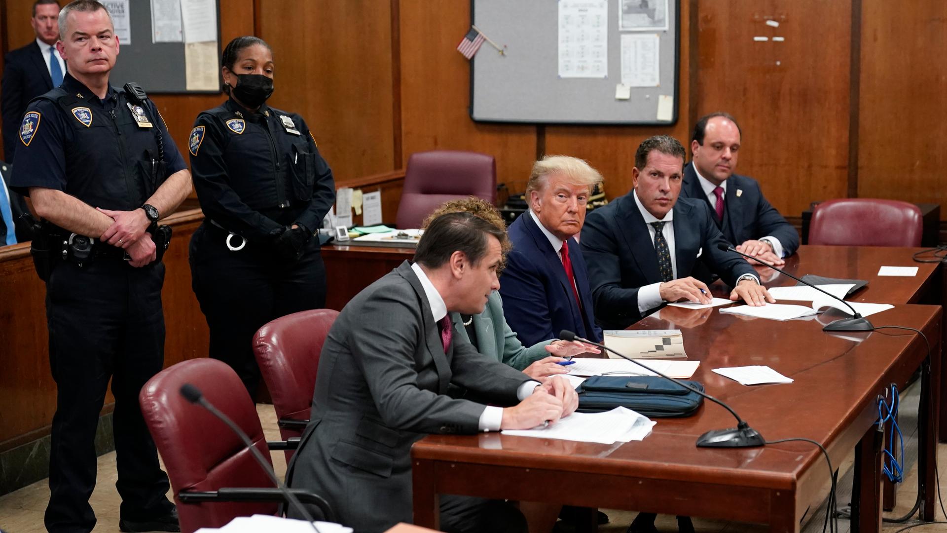 Donald Trump sitzt auf der Anklagebank eines Gerichts; links und rechts von ihm je zwei Anwälte. Im Hintergrund stehen uniformierte Beamte.