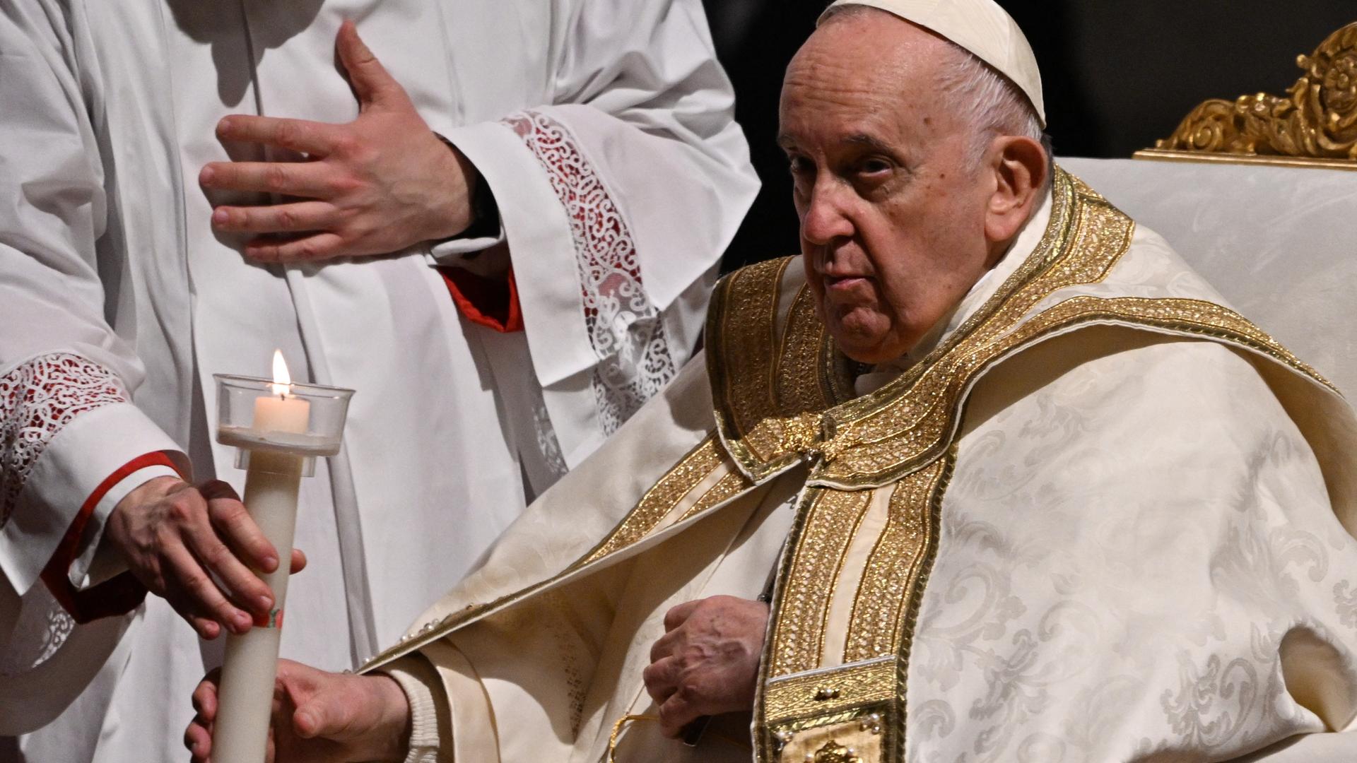 Der Papst sitzt in einem Sessel und bekommt eine Kerze gereicht.