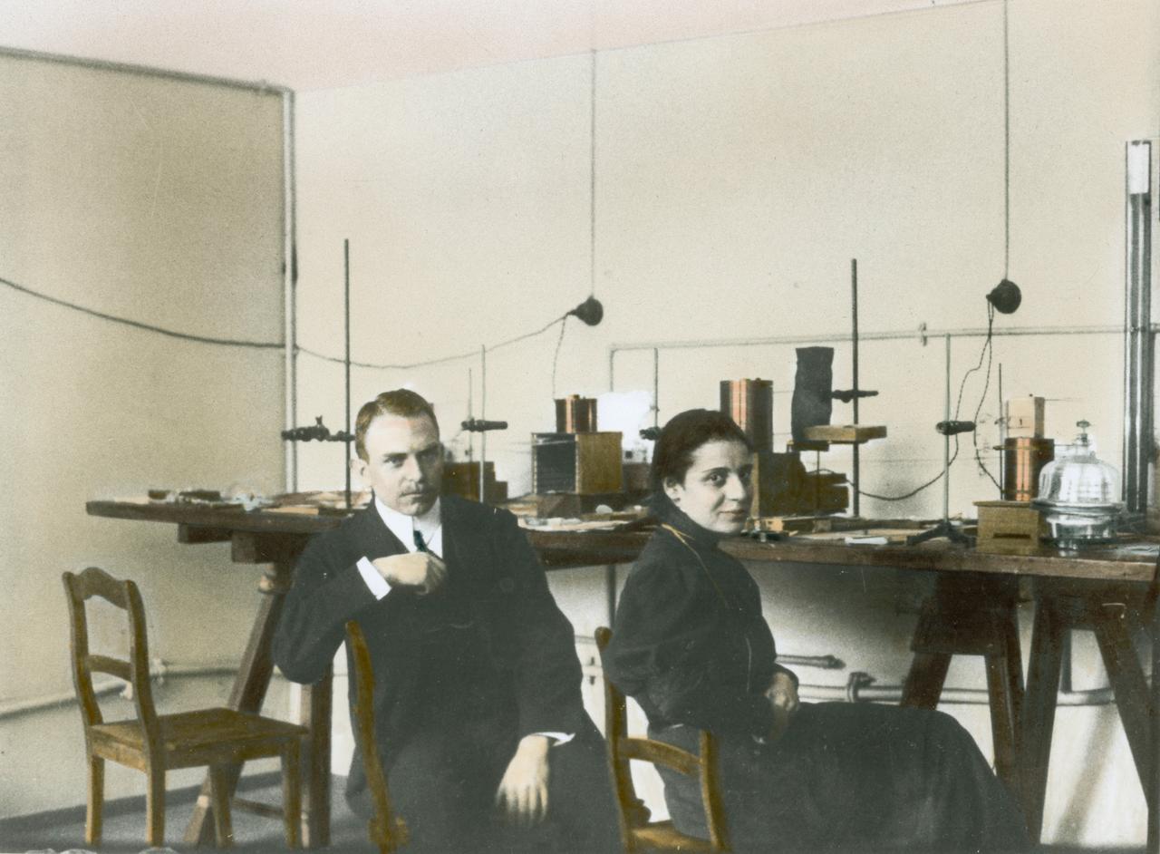 Nachkolorierte Aufnahme von Otto Hahn mit Lise Meitner sitzend im Labor um 1910.