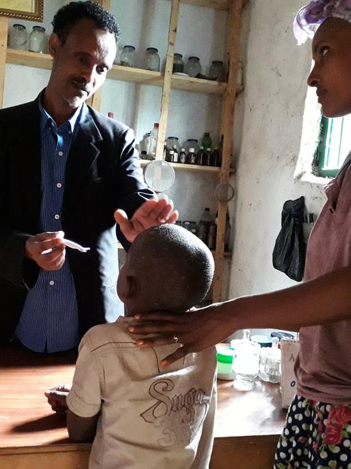 Der Heiler Tegistu behandelt in Äthiopien einen fünfjährigen Jungen, der an einer Pilzerkrankung leidet. Er hat seine Hand auf dem Kopf des Jungen. Daneben steht dessen Mutter. Im Hintergrund sind Gläser mit Pflanzen.