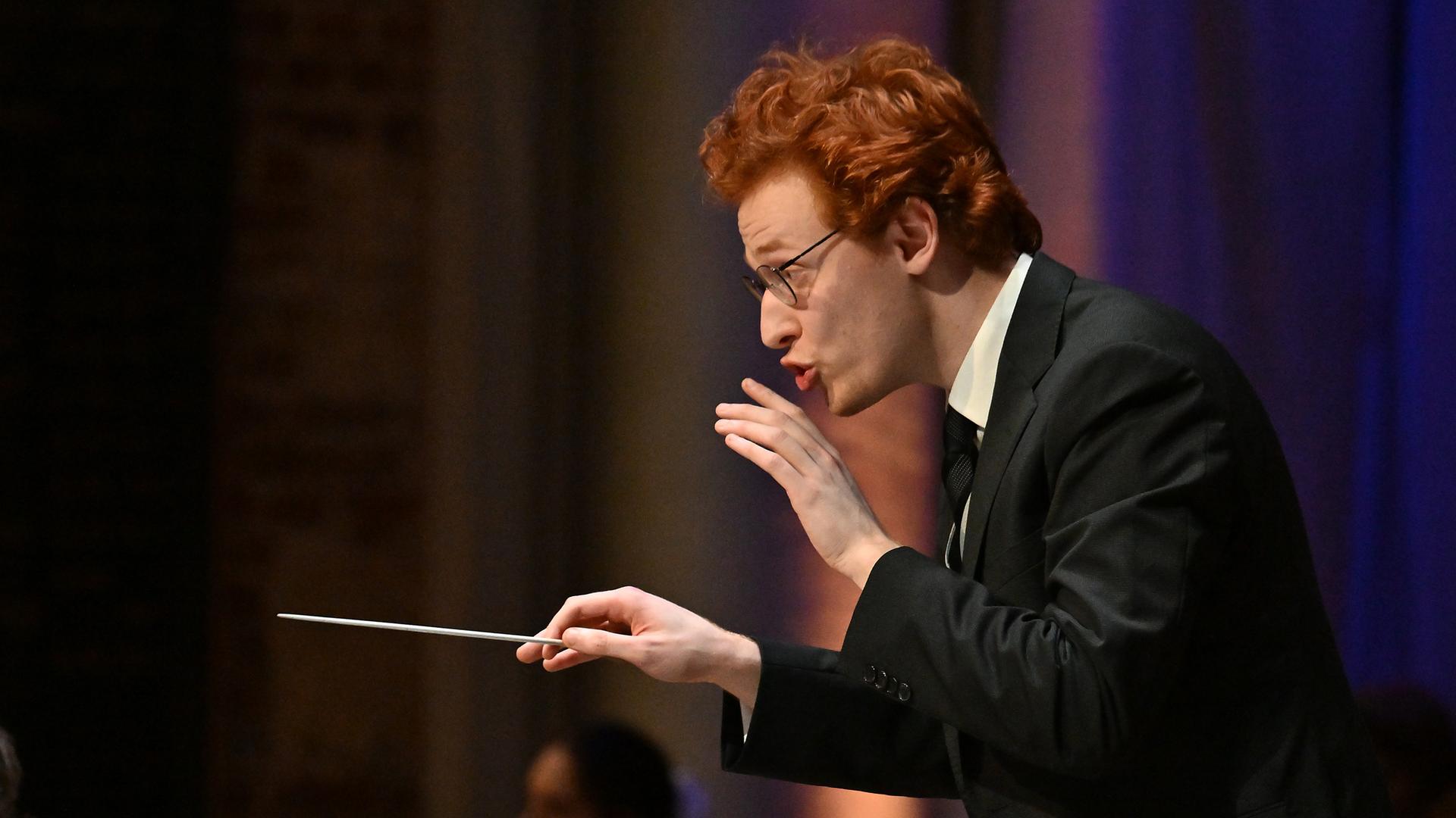 Ein junger Mann mit Brille und roten kurzen Haaren gibt ein Zeichen, dass das Orchester leiser spielen soll. In der rechten Hand hält er einen Dirigierstab.