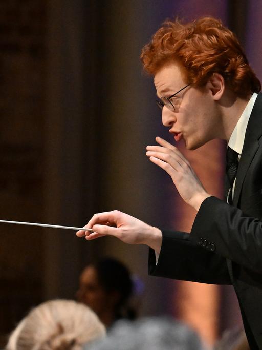 Ein junger Mann mit Brille und roten kurzen Haaren gibt ein Zeichen, dass das Orchester leiser spielen soll. In der rechten Hand hält er einen Dirigierstab.