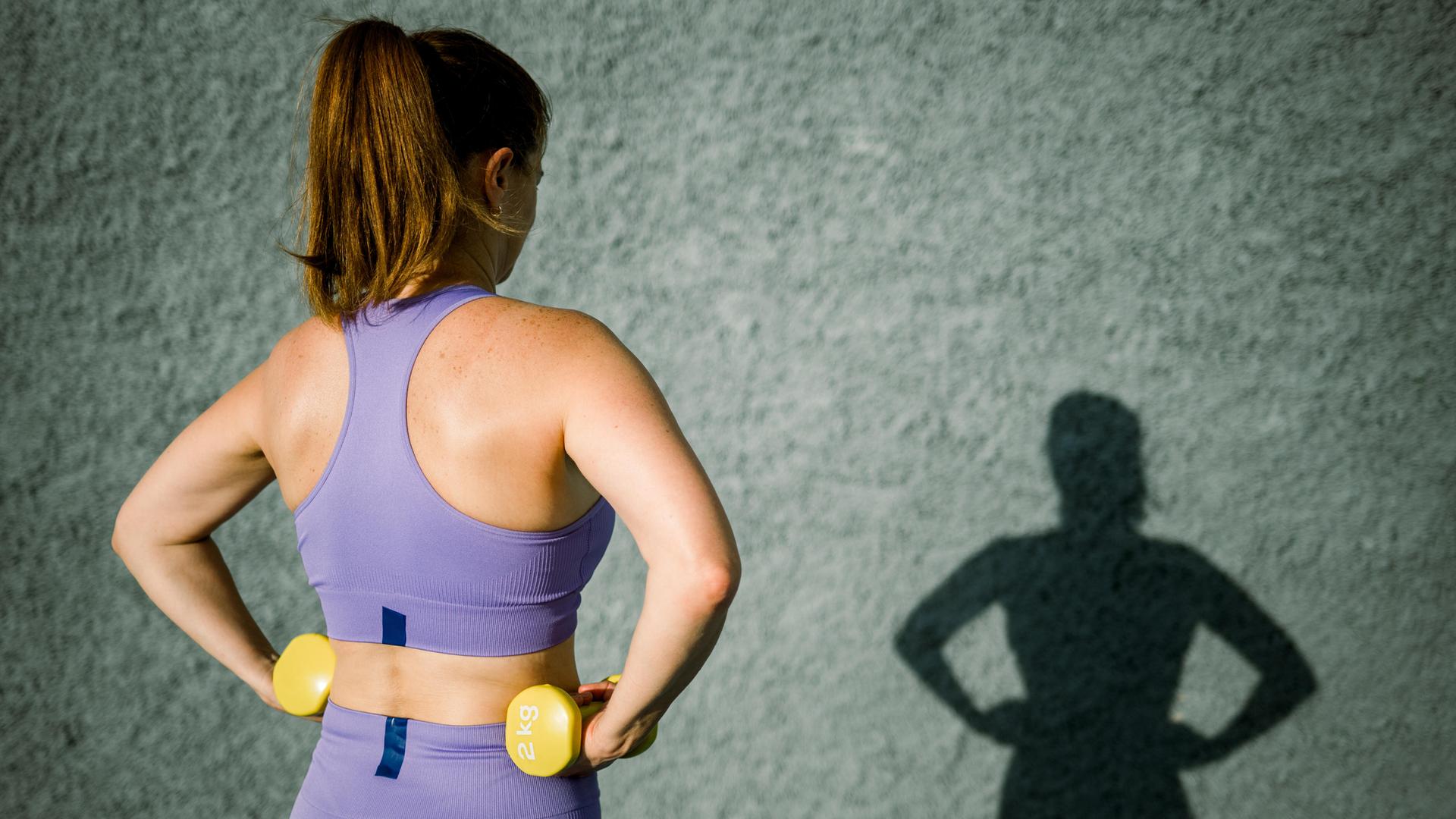 Eine Frau in violetten Sportklamotten von hinten, sie hat zwei gelbe Hanteln in jeder Hand, auch ihr Schatten ist zu sehen