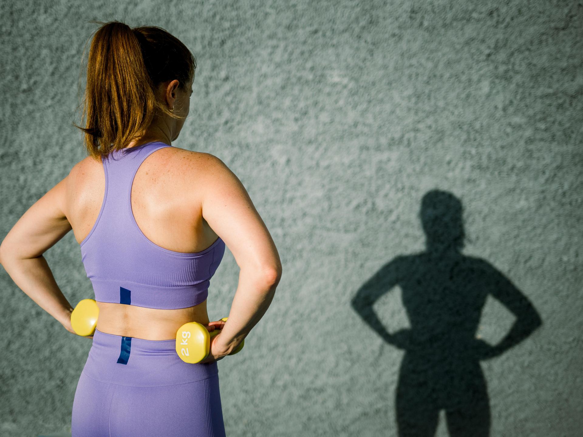 Eine Frau in violetten Sportklamotten von hinten, sie hat zwei gelbe Hanteln in jeder Hand, auch ihr Schatten ist zu sehen