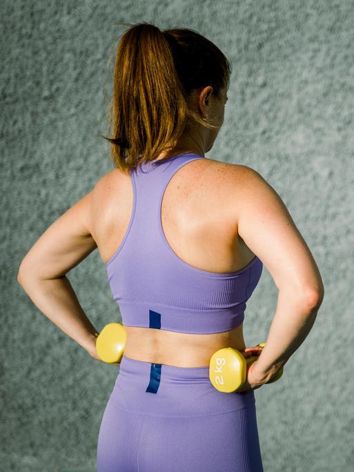 Eine Frau in violetten Sportklamotten von hinten, sie hat zwei gelbe Hanteln in jeder Hand, auch ihr Schatten ist zu sehen Eine Frau in violetten Sportklamotten von hinten, sie hat zwei gelbe Hanteln in jeder Hand, auch ihr Schatten ist zu sehen