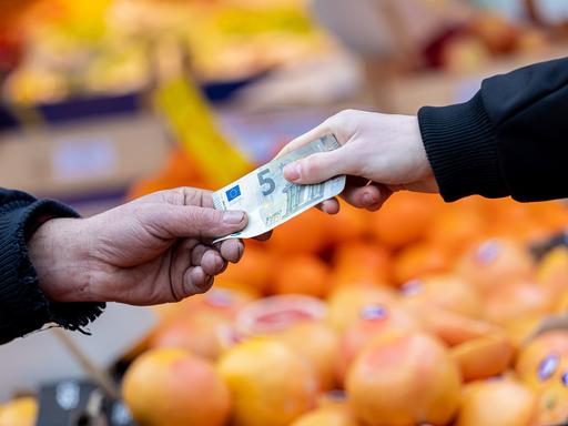 Ein Kunde bezahlt seinen Einkauf an einem Stand auf einem Wochenmarkt mit einem Fünf-Euro-Schein.
