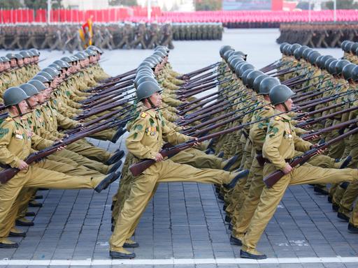 Die Soldaten gehen in mehreren Reihen hintereinander im Stechschritt über einen Platz. Sie tragen Gewehre. 