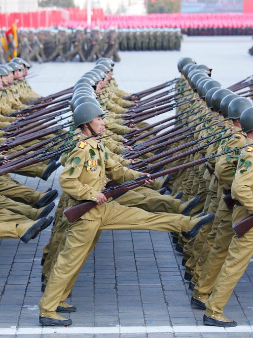 Die Soldaten gehen in mehreren Reihen hintereinander im Stechschritt über einen Platz. Sie tragen Gewehre. 