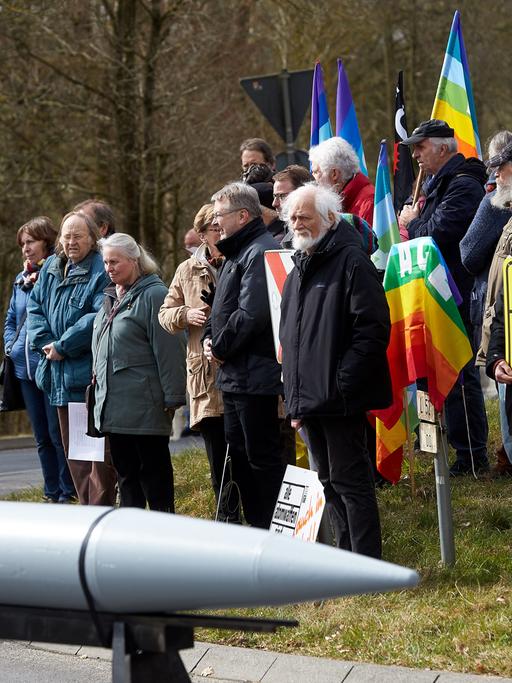 Zum Auftakt einer Friedensaktion im März 2018 am Fliegerhorst Büchel haben sich Demonstranten an dem Fliegerhosrt versammelt. Im Vordergrund ein schematisch nachgebaute Rakete. Im Hintergrund stehen Menschen, zum Teil halten sie Friedensymbole wie die Regenbogenfahne.
