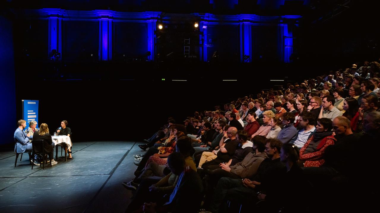 Bei der Aufzeichnung des Politikpodcasts am Neuen Theater in Halle (Saale) diskutieren Korrespondentinnen an einem Tisch, das Publikum lauscht von ansteigenden Sitzreihen.