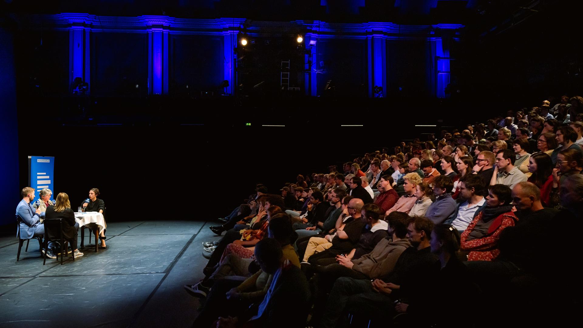 Bei der Aufzeichnung des Politikpodcasts am neuen theater in Halle (Saale) diskutieren Korrespondentinnen an einem Tisch, das Publikum lauscht aus ansteigenden Sitzreihen. Bei der Aufzeichnung des Politikpodcasts am neuen theater in Halle (Saale) diskutieren Korrespondentinnen an einem Tisch, das Publikum lauscht aus ansteigenden Sitzreihen.