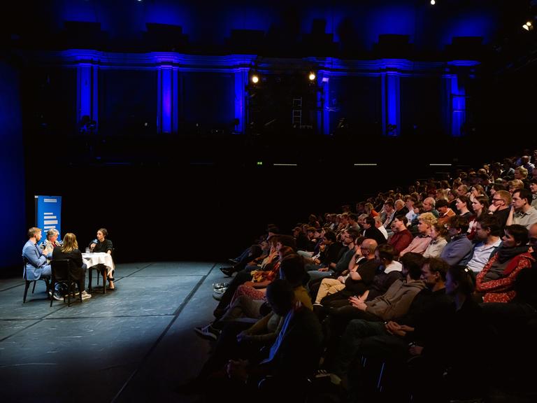 Bei der Aufzeichnung des Politikpodcasts am Neuen Theater in Halle (Saale) diskutieren Korrespondentinnen an einem Tisch, das Publikum lauscht von ansteigenden Sitzreihen. Bei der Aufzeichnung des Politikpodcasts am Neuen Theater in Halle (Saale) diskutieren Korrespondentinnen an einem Tisch, das Publikum lauscht von ansteigenden Sitzreihen.
