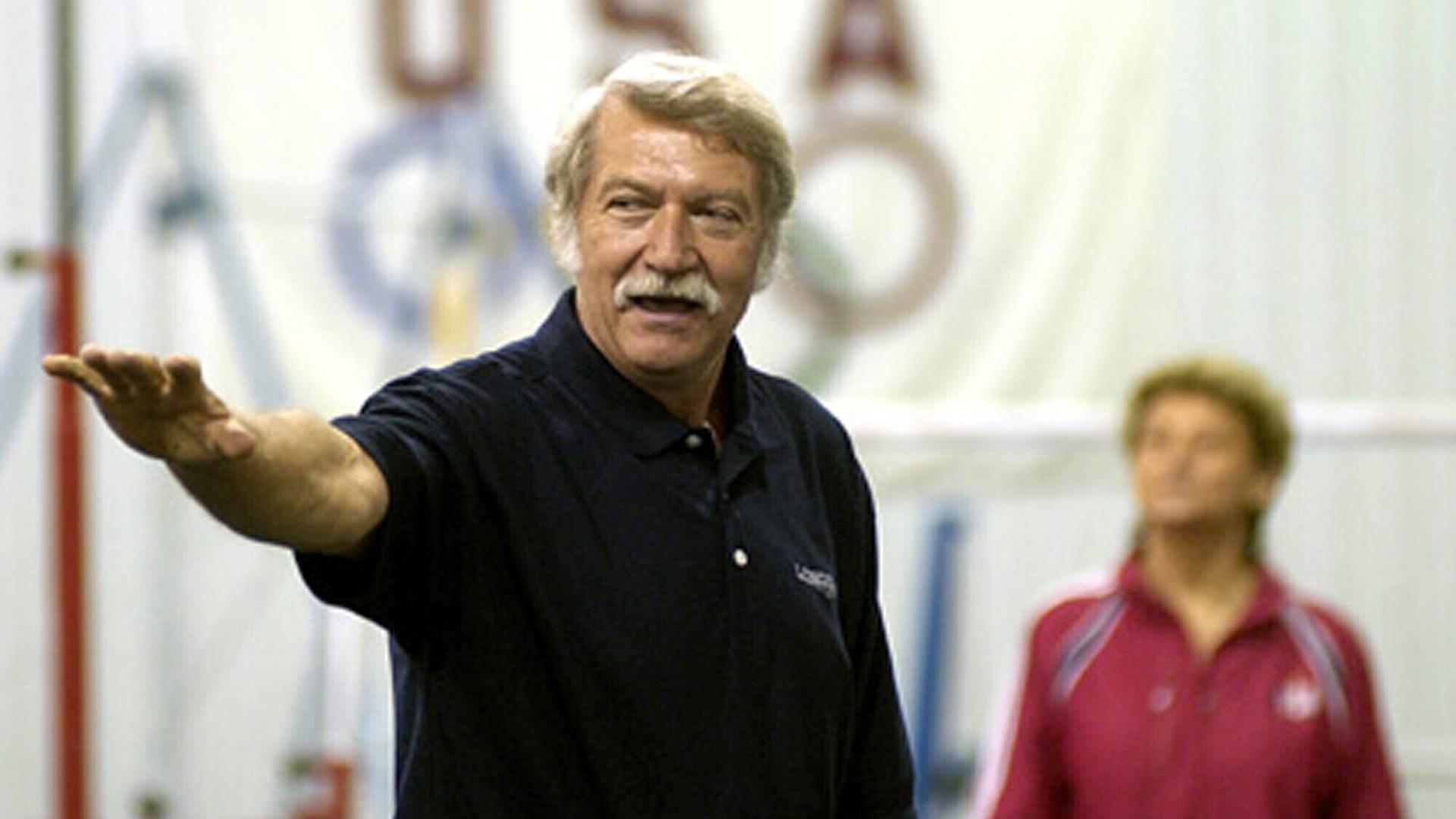 Béla Károlyi steht in der Turnhalle und zeigt auf etwas.