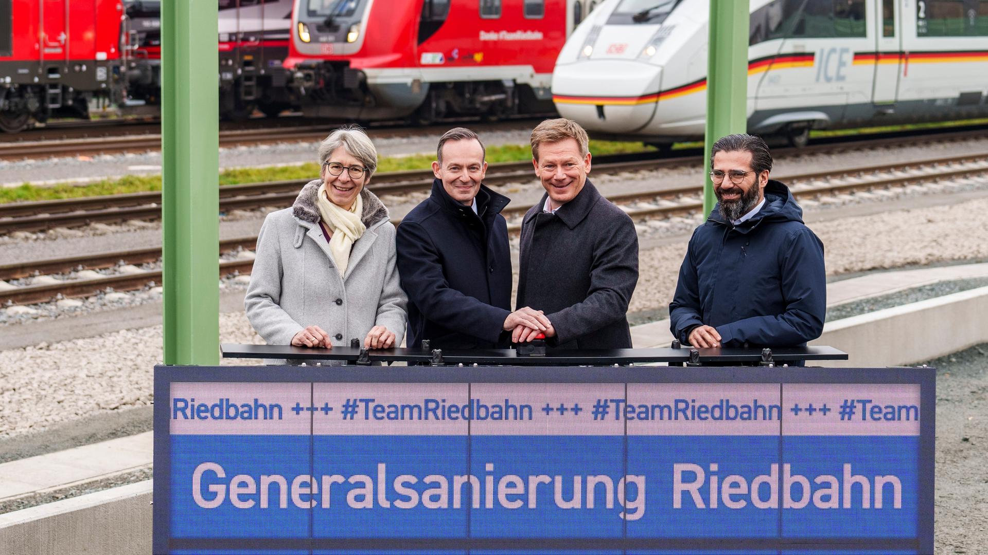 Die beiden stehen zwischen zwei Landespolitikern auf einem Bahnsteig. Wissing und Lutz schütteln sich die Hände. Sie stehen vor einer Anzeige, auf der "Generalsanierung Riedbahn" steht. Im Hintergrund drei Züge. 