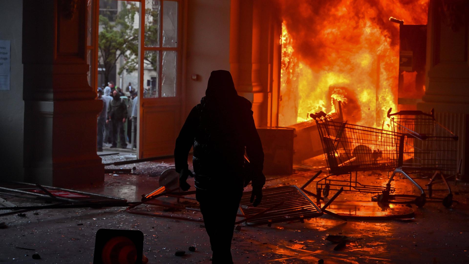 Die Silhouette einer in schwarz gekleideten Personen mit Kapuzenjacke vor den Flammen des Rathauses von Limoges. Das Gesicht der Person ist nicht erkennbar. 