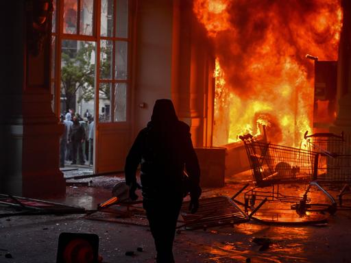 Die Silhouette einer in schwarz gekleideten Personen mit Kapuzenjacke vor den Flammen des Rathauses von Limoges. Das Gesicht der Person ist nicht erkennbar. Die Silhouette einer in schwarz gekleideten Personen mit Kapuzenjacke vor den Flammen des Rathauses von Limoges. Das Gesicht der Person ist nicht erkennbar.
