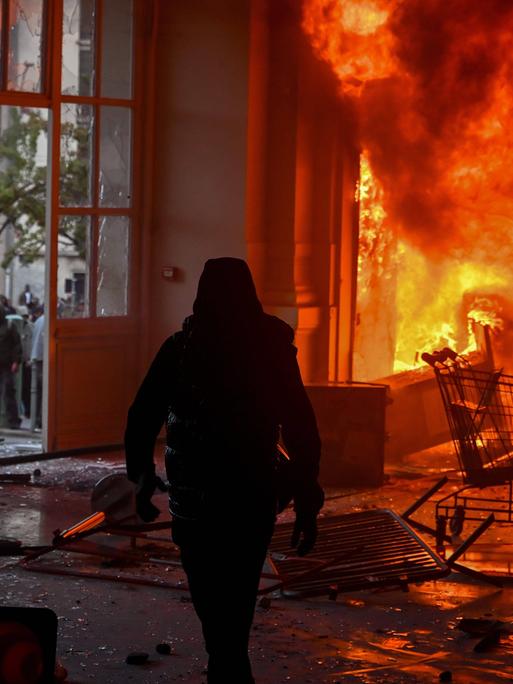 Die Silhouette einer in schwarz gekleideten Personen mit Kapuzenjacke vor den Flammen des Rathauses von Limoges. Das Gesicht der Person ist nicht erkennbar. Die Silhouette einer in schwarz gekleideten Personen mit Kapuzenjacke vor den Flammen des Rathauses von Limoges. Das Gesicht der Person ist nicht erkennbar.