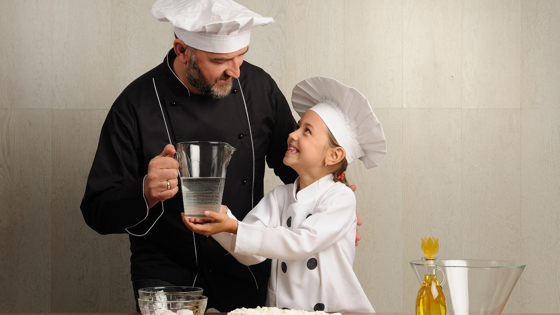 Ein Vater und seine Tochter beim Kochen, beide tragen weiße Kochmützen und hantieren mit Öl und Wasser, im Vordergrnd sieht man eine Schüssel und Mehl