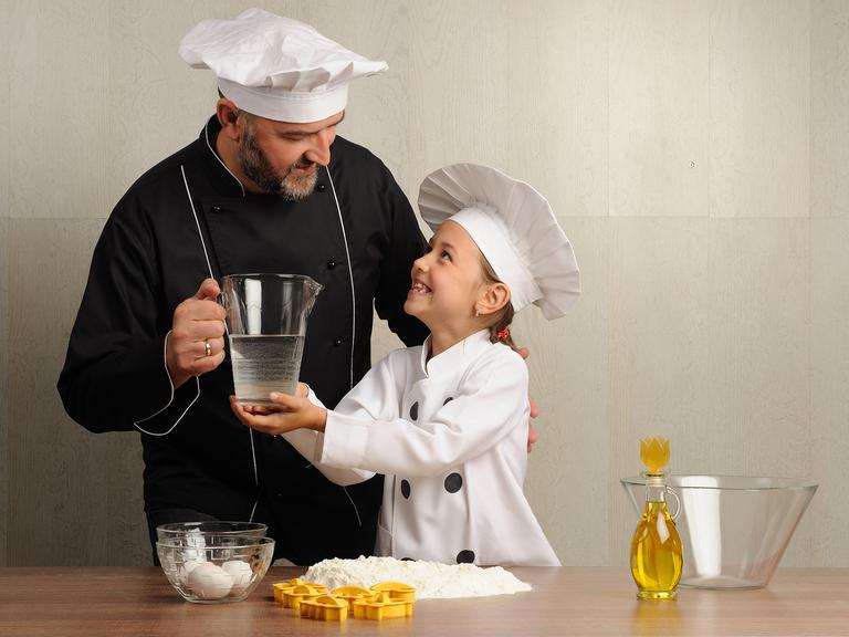 Ein Vater und seine Tochter beim Kochen, beide tragen weiße Kochmützen und hantieren mit Öl und Wasser, im Vordergrnd sieht man eine Schüssel und Mehl
