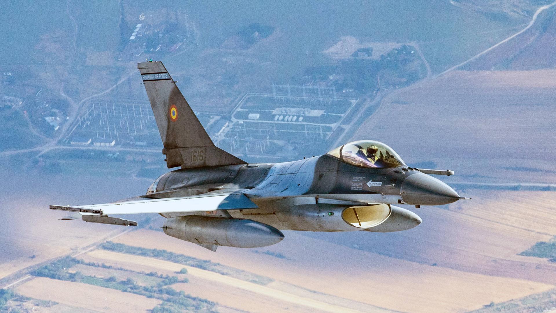 Ein F-16-Kampf-Flugzeug fliegt hoch über Wiesen und Feldern.