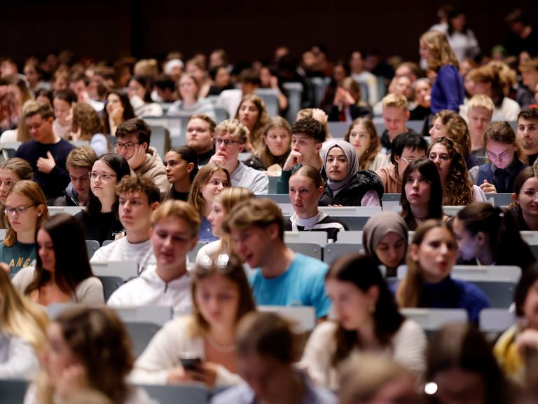 In einem großen Hörsaal sitzen viele Studierenden in den Reihen. Es sind Erstsemester bei einer Einführungsvorlesung.