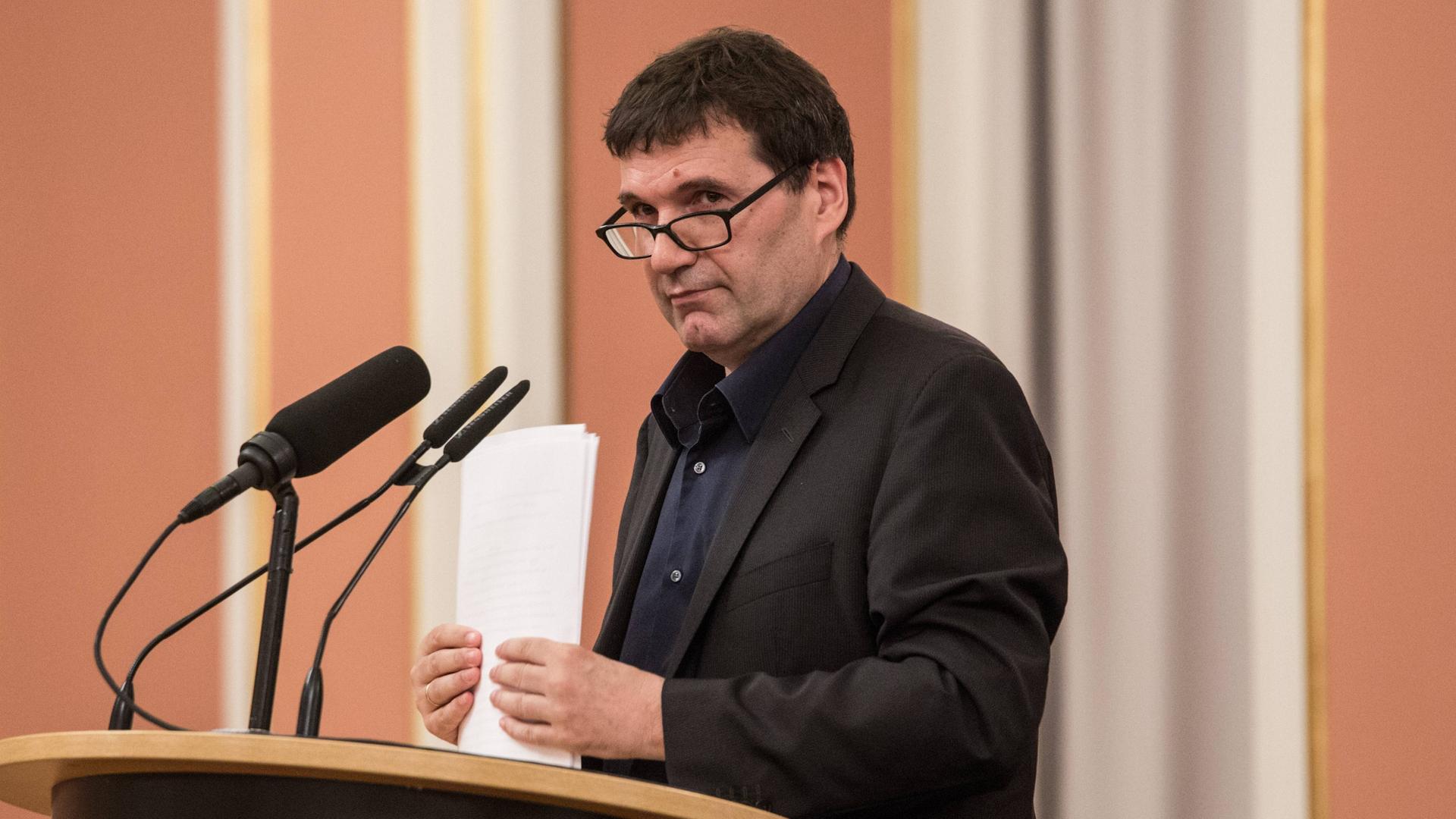 Der Büchner-Preisträger Oswald Egger steht an einem Rednerpult vor Mikrofonen und hält Papier in der Hand.