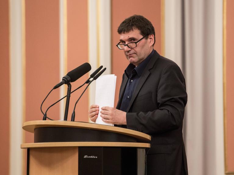 Der Büchner-Preisträger Oswald Egger steht an einem Rednerpult vor Mikrofonen und hält Papier in der Hand.