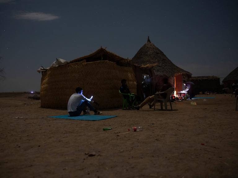 Ein Geflüchteter aus der äthiopischen Konfliktregion Tigray sitzt in der Nacht vor einer Lehmhütte und blickt auf das hell erleuchtete Display seines Mobiltelefons.
