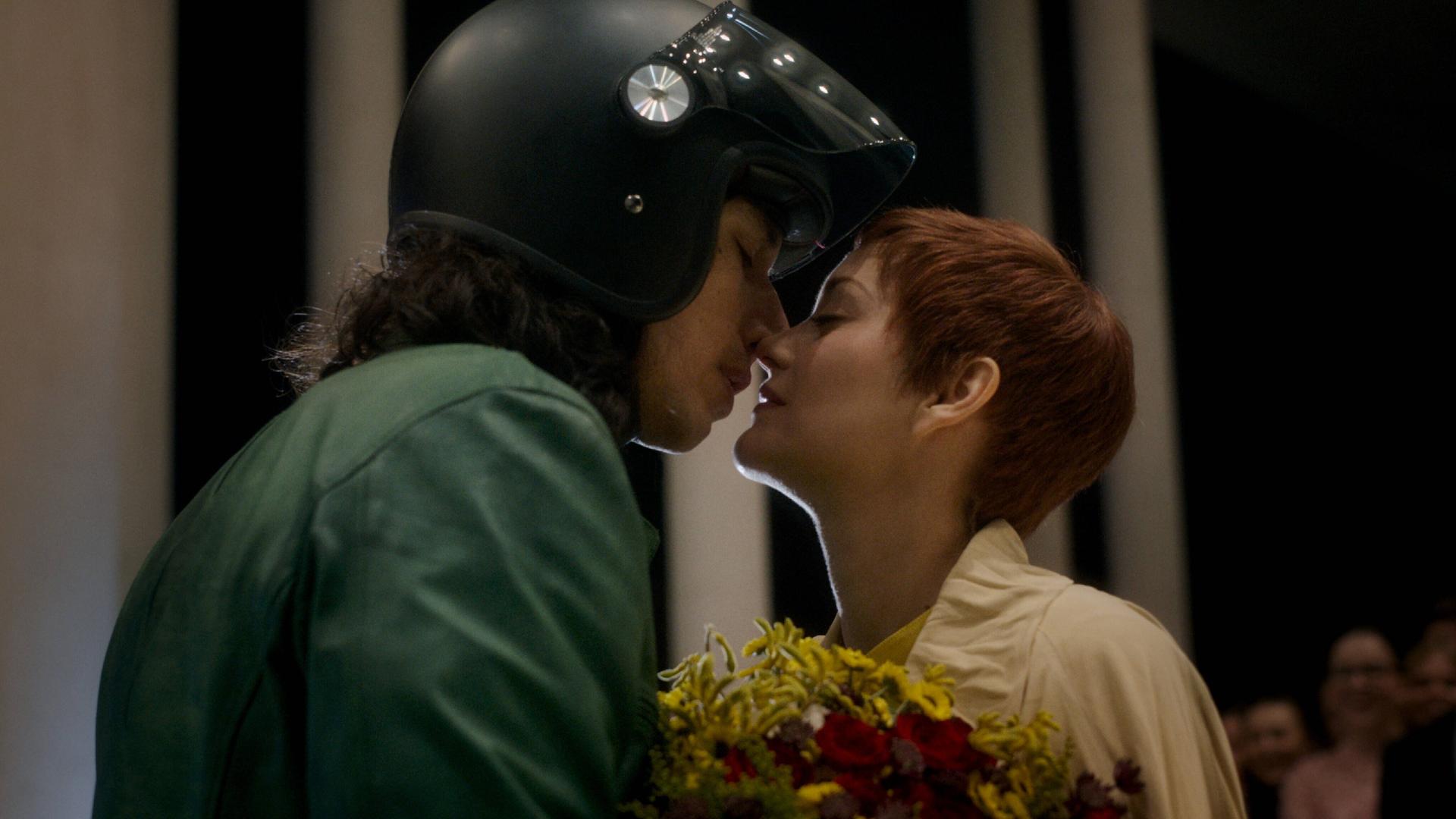 Ein Mann mit Motorradhelm küsst eine Frau, die einen Blumenstrauß hält.