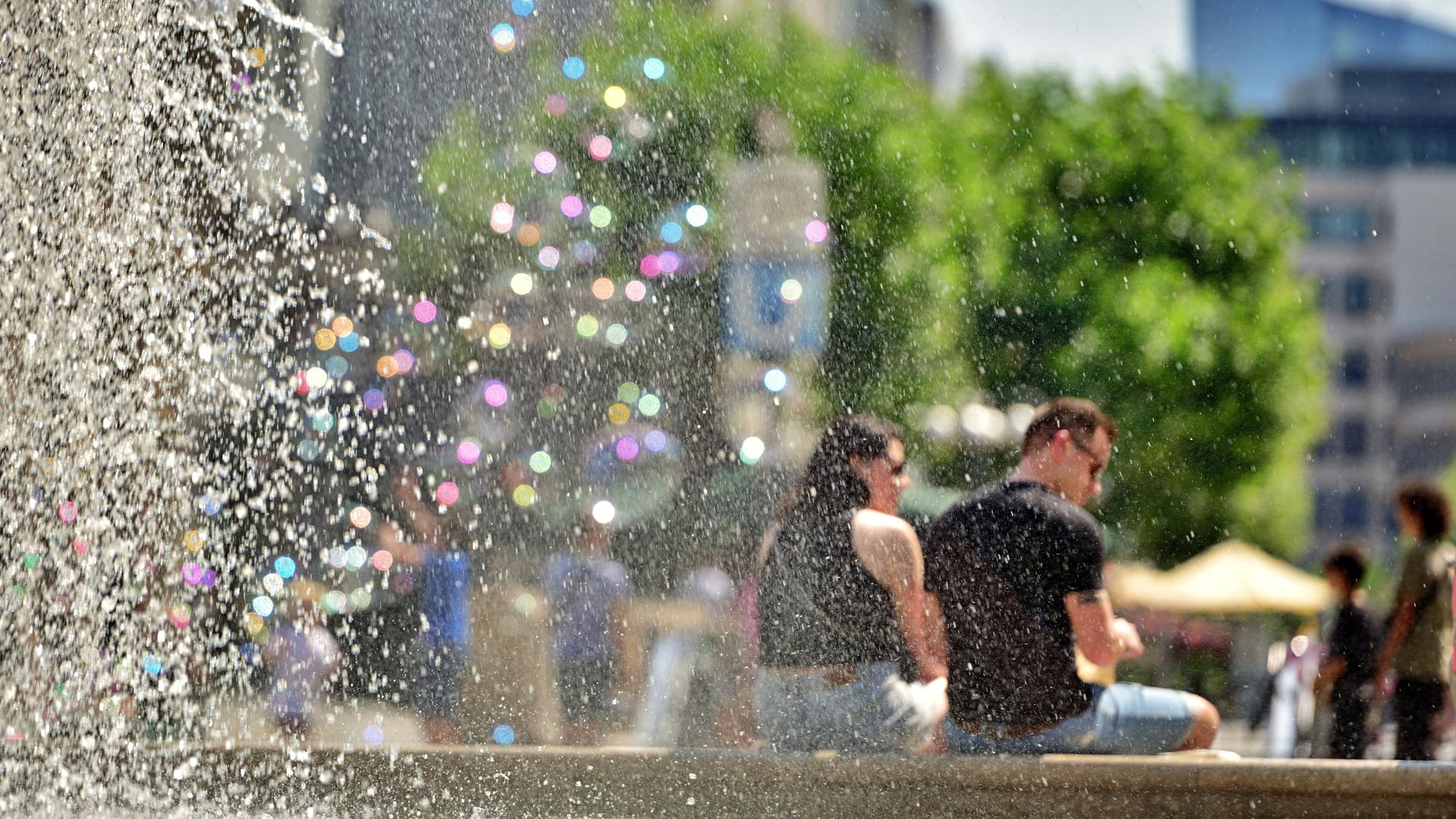 Am großen Brunnen vor der alten Oper in Frankfurt sitzen Menschen und trotzen der Hitze. Links im Bild sind Wassertropfen in bunten Pastelltönen zu sehen, rechts sitzt ein Pärchen am Brunnenrand. Am großen Brunnen vor der alten Oper in Frankfurt sitzen Menschen und trotzen der Hitze. Links im Bild sind Wassertropfen in bunten Pastelltönen zu sehen, rechts sitzt ein Pärchen am Brunnenrand.