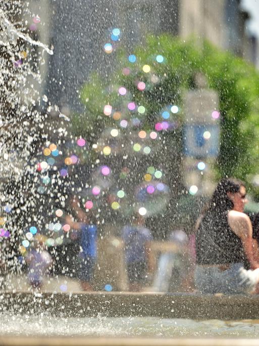 Am großen Brunnen vor der alten Oper in Frankfurt sitzen Menschen und trotzen der Hitze. Links im Bild sind Wassertropfen in bunten Pastelltönen zu sehen, rechts sitzt ein Pärchen am Brunnenrand. 