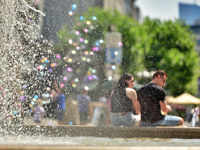 Am großen Brunnen vor der alten Oper in Frankfurt sitzen Menschen und trotzen der Hitze. Links im Bild sind Wassertropfen in bunten Pastelltönen zu sehen, rechts sitzt ein Pärchen am Brunnenrand. 