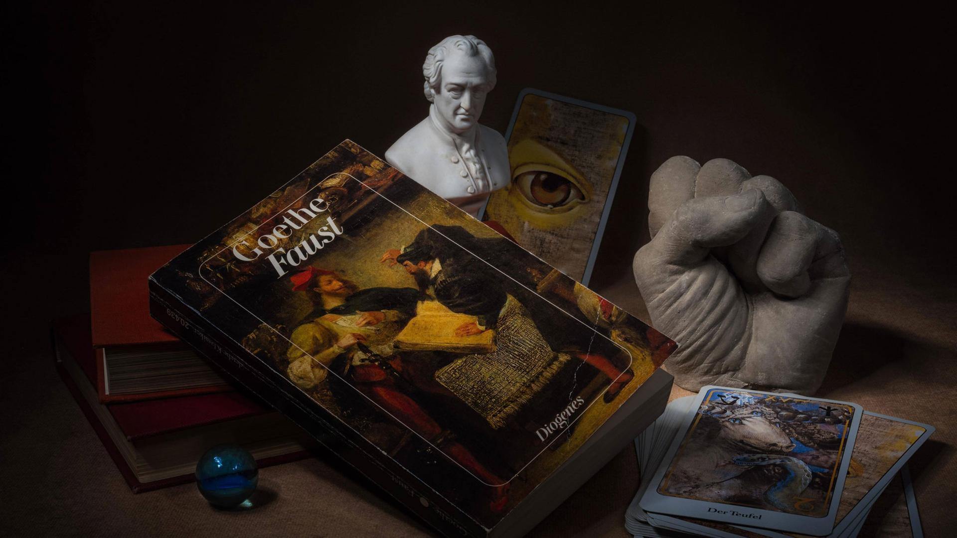 Hörspielkomödie über Goethes "Faust" und die Mühen des Deutschwerdens. Zu sehen: Stilleben mit Büchern, Glaskugel, Tarotkarten, Goethe-Büste, das Buch "Faust" und einer Plastikhand zu einer Faust geballt. 