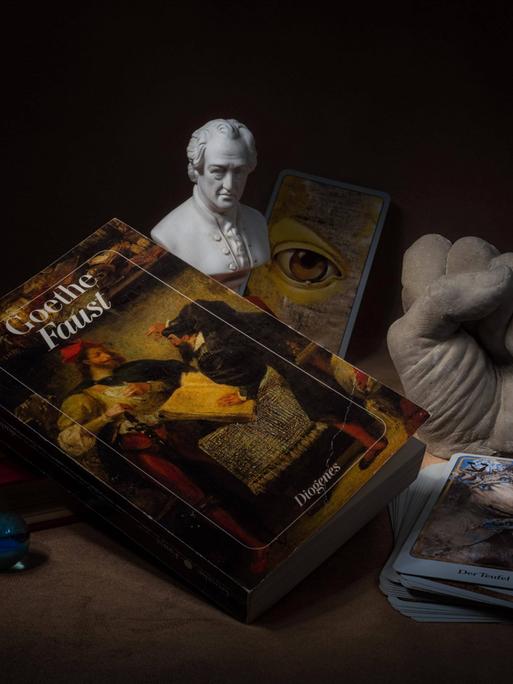Hörspielkomödie über Goethes "Faust" und die Mühen des Deutschwerdens. Zu sehen: Stilleben mit Büchern, Glaskugel, Tarotkarten, Goethe-Büste, das Buch "Faust" und einer Plastikhand zu einer Faust geballt. 