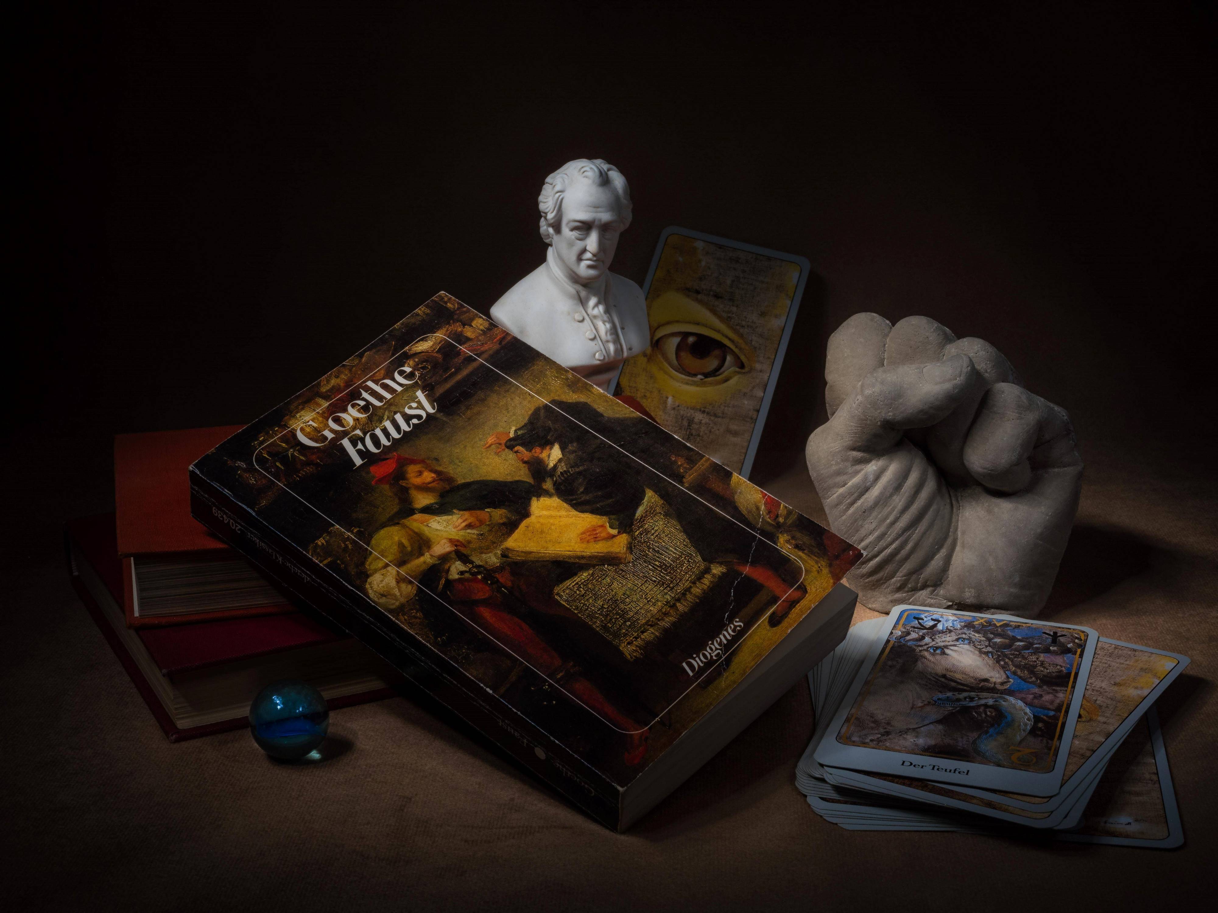 Hörspielkomödie über Goethes "Faust" und die Mühen des Deutschwerdens. Zu sehen: Stilleben mit Büchern, Glaskugel, Tarotkarten, Goethe-Büste, das Buch "Faust" und einer Plastikhand zu einer Faust geballt. 