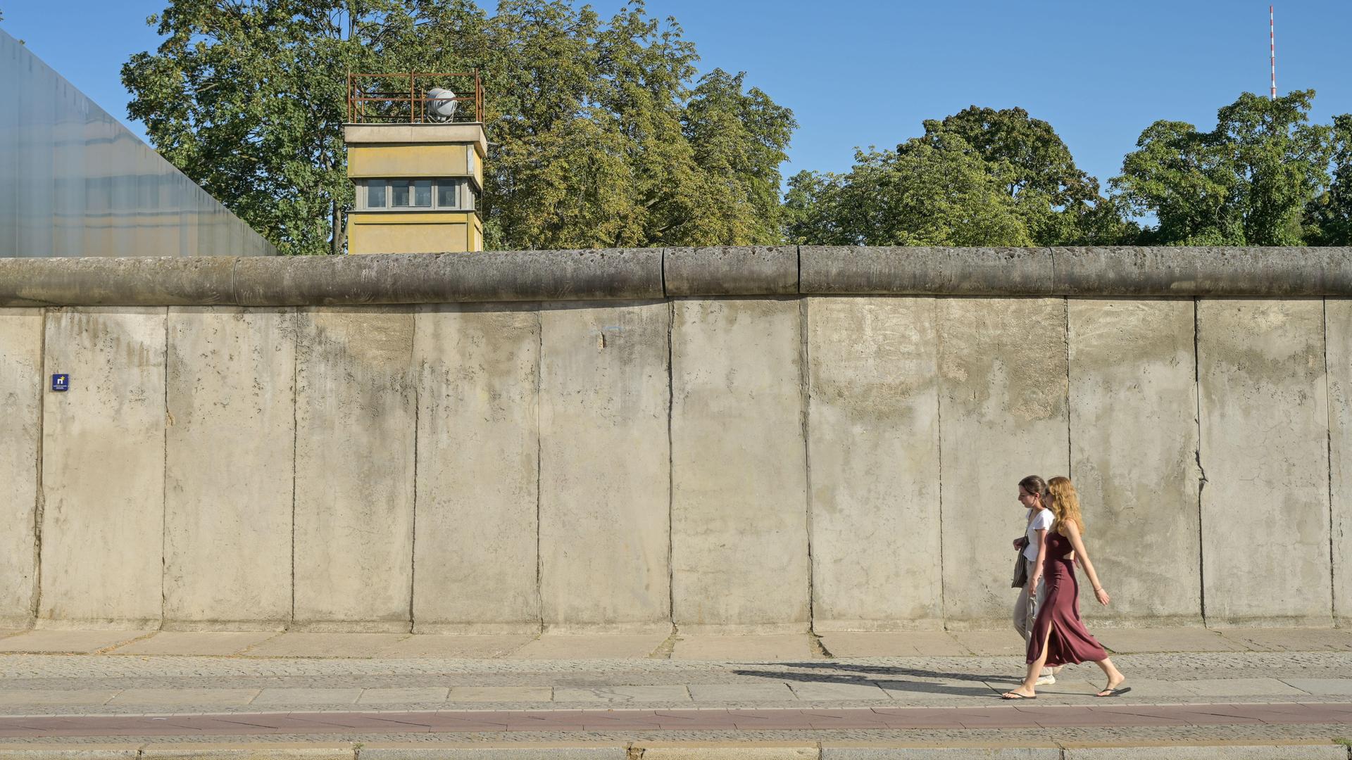 Blick auf die Berliner Mauer, hinter der Mauer ist ein Wachturm zu sehen. Davor laufen zwei Personen entlang.