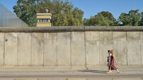 Blick auf die Berliner Mauer, hinter der Mauer ist ein Wachturm zu sehen. Davor laufen zwei Personen entlang.