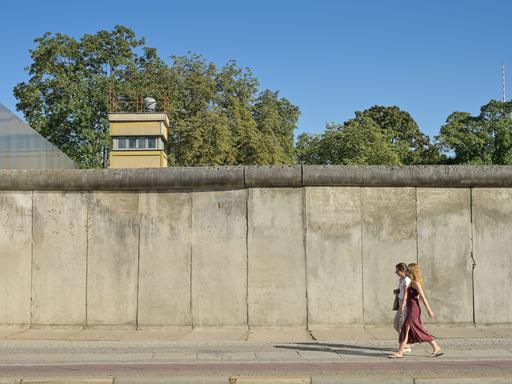 Blick auf die Berliner Mauer, hinter der Mauer ist ein Wachturm zu sehen. Davor laufen zwei Personen entlang.