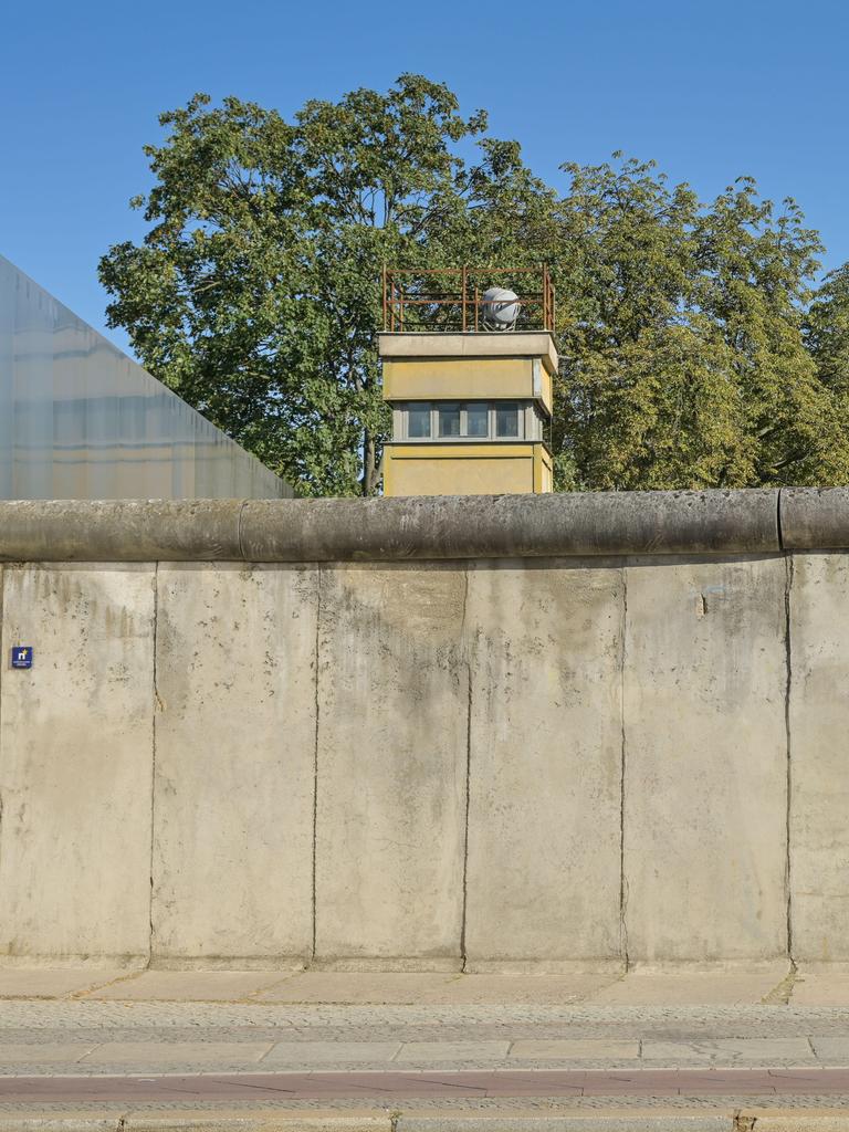 Blick auf die Berliner Mauer, hinter der Mauer ist ein Wachturm zu sehen. Davor laufen zwei Personen entlang.