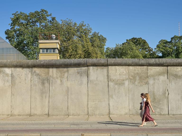 Blick auf die Berliner Mauer, hinter der Mauer ist ein Wachturm zu sehen. Davor laufen zwei Personen entlang. Blick auf die Berliner Mauer, hinter der Mauer ist ein Wachturm zu sehen. Davor laufen zwei Personen entlang.